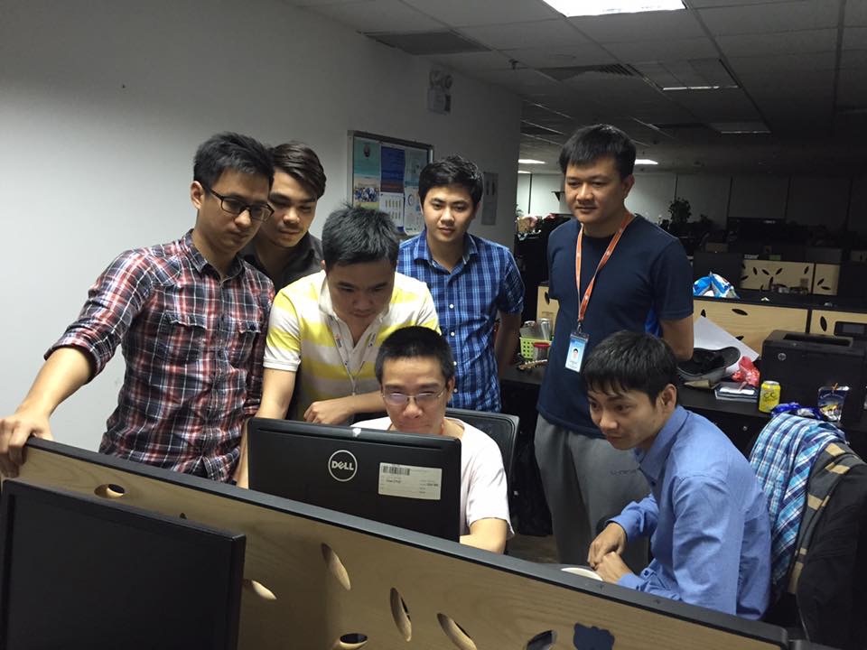 Bảy người quanh một màn hình: một người ngồi code, sáu người đứng phía sau xem